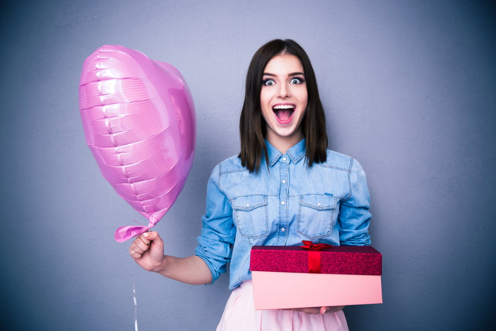 Amazed woman holding balloon and gift box over gray background. Looking at camera.